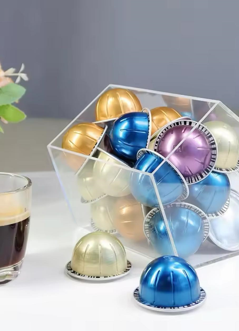 Colorful coffee capsules in a transparent holder with a glass of coffee on a white surface.